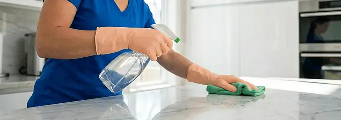 Maid service worker spraying and wiping a kitchen countertop with a microfiber cloth for sanitizing, Rochester, NY.